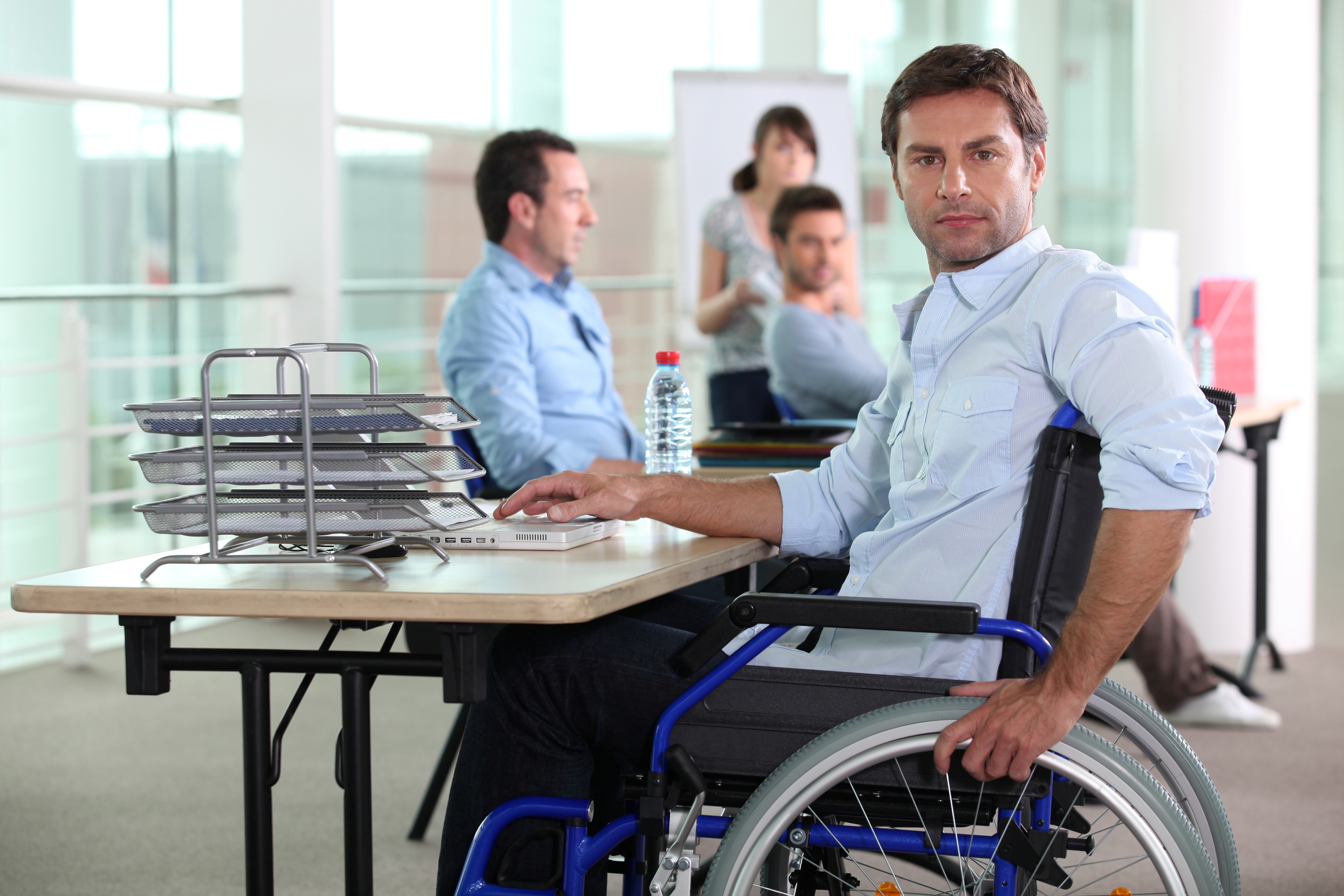 Image shows a man in a wheelchair, at a desk with his laptop, papers, and co-workers.