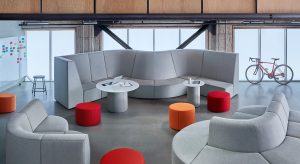 Images shows industrial style workplace with glass sliding doors. At first glance you see a large seating area with a high back bench seat that twist and turns at the centerpiece. Additional lower back seating is positioned across from it with pops of color proivded by the ottomans that double as additional seating. On the far left wall you can see a white board hung to provide the team a space to brainstorm ideas.